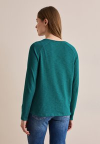 Tummansinihkeä pitkähihainen paita pyöreällä pääntiellä ja raglan-hihoilla, yhdistettynä sinisiin farkkuihin. Pehmeä kangas, minimalistinen muotoilu.