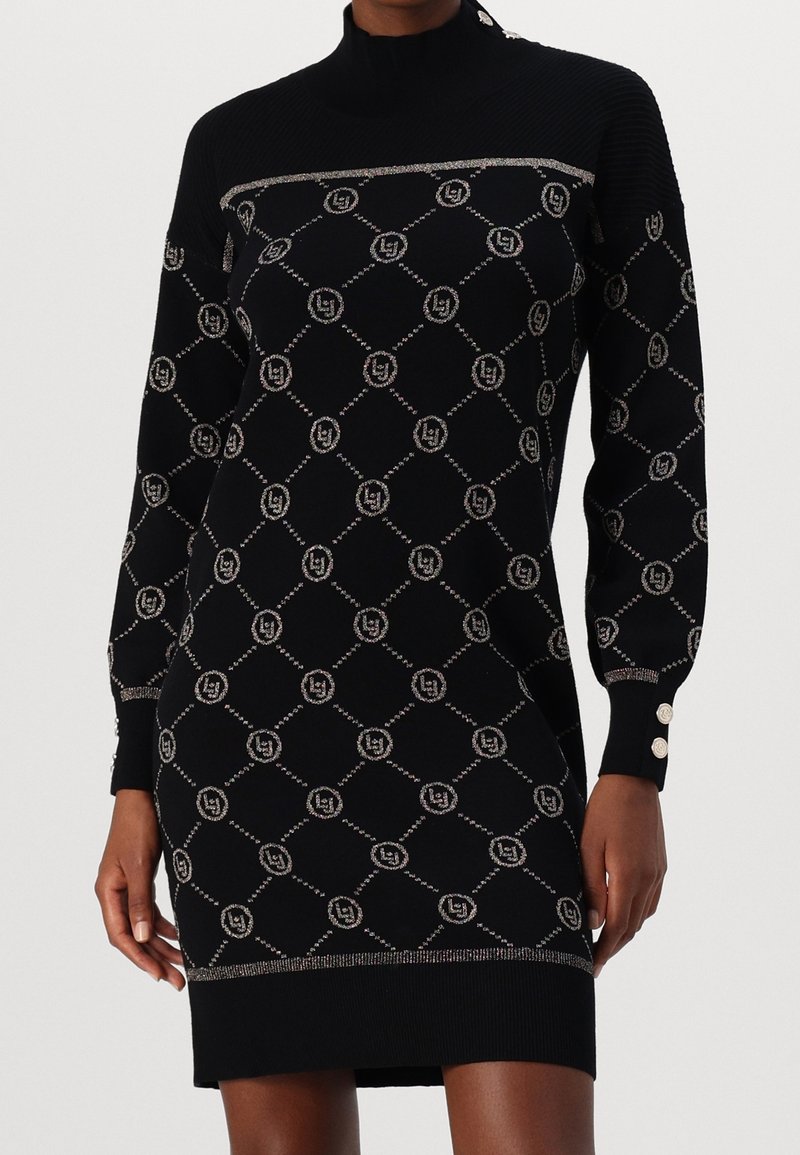 Robe longue noire en tricot avec motif argenté en losange et cercle, col montant, détails de boutons aux poignets, portée par une personne debout.