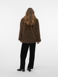 Moteris su ilgais banguotais plaukais, vilkinti rudą kailinį paltą, juodus aksominius kelnius ir juodas aukštakulnes batus, stovi prieš baltą foną.