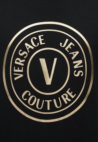 Χρυσλό κυκλικό λογότυπο Versace Jeans Couture με ένα μεγάλο "V" στο κέντρο σε μαύρο φόντο.