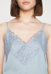 Ljusblå topp med spetsdetaljer längs V-ringningen och axlarna. Tyget ser slätt och något glänsande ut, med en delikat textur.