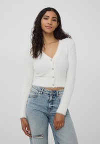 Cardigan bianco cropped realizzato in materiale soffice, con scollatura a V profonda, maniche lunghe e cinque bottoni luminosi.