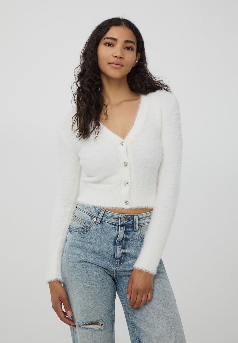 Cardigan bianco cropped realizzato in materiale soffice, con scollatura a V profonda, maniche lunghe e cinque bottoni luminosi.