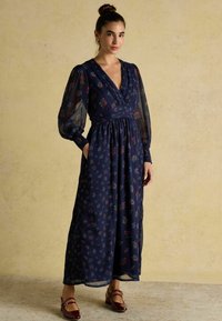 Tume lilleline maksimekko, millel on sügav V-kaelus, pikad läbipaistvad puhv-sleeved, kogutud vöökoht ja küljetaskud; kombineeritud burgundi värvi kingadega.