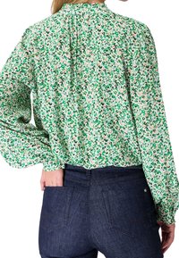 Blusa de leopardo en verde y blanco con mangas largas, detalle plisado en la parte trasera y un cuello alto, combinada con jeans de mezclilla oscura.