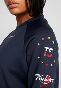 Sudadera de color marino con un cuello redondo acanalado, puños ajustados y gráficos de logotipo vibrantes en rojo, blanco y amarillo en la manga.