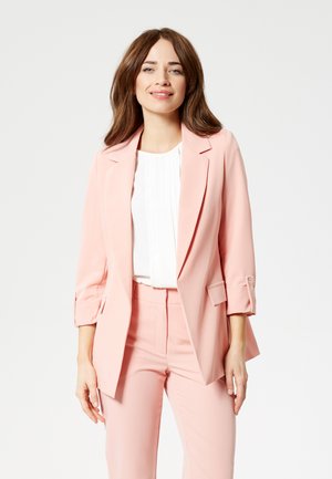Donna con capelli castani che indossa un blazer rosa chiaro e pantaloni abbinati sopra una camicetta bianca, sorridente su uno sfondo chiaro e uniforme.