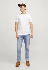 Bela bombažna majica, svetlo modre kavbojke in rjavi škornji. Model nosi rjavo kapo in stoji na nevtralnem ozadju.