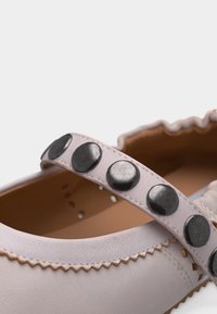 Κοντινή εικόνα λουριού παπουτσιού από δέρμα ανοιχτού ροζ χρώματος με μεγάλα στρογγυλά μαύρα καρφιά και διακοσμητικό κυματιστό περίγραμμα στο επάνω μέρος του παπουτσιού.