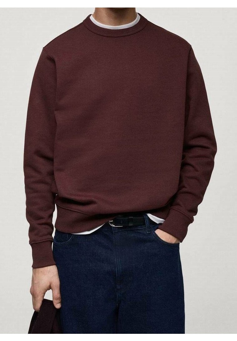 Persoon draagt een kastanjebruine sweater met ronde hals over een wit shirt en donkerblauwe spijkerbroek, één hand in de zak en de andere houdt een stuk stof vast.