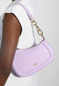 Sac à main en cuir lavande avec une silhouette incurvée, doté d'une bandoulière en chaîne dorée et d'une fermeture éclair. Accent logo doré à l'avant.