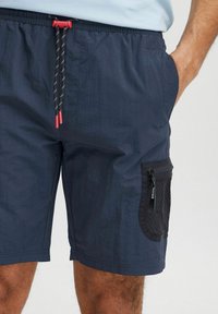 Shorts de baie bleu marin dintr-un material ușor, cu un brâu elastic și accente roșii, având un detaliu de buzunar din plasă pe lateral.