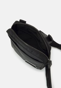 Borsa a tracolla nera in nylon con chiusura a zip, dotata di scomparti multipli e una superficie liscia. Include un'elegante etichetta del marchio e una tracolla regolabile.