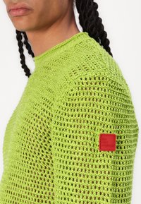 Suéter verde tricotado com um design em renda, decote redondo e um logo vermelho na manga esquerda.