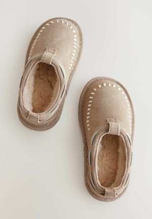 Beige Wildlederhausschuhe mit einem weichen Futter aus Fell, ausgestattet mit weißem Dekostich und einer strukturierten Gummisohle. Rutschfestes Design ohne Verschlüsse.