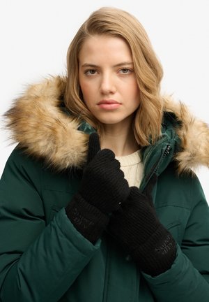 Jeune femme portant des gants noirs et un parka vert avec une capuche bordée de fourrure, face à l'avant sur un fond uni.
