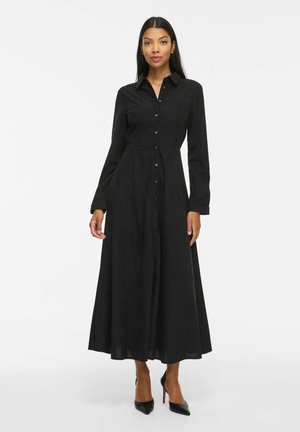 Femme debout portant une longue robe noire boutonnée à manches longues et des chaussures à talons noirs sur un fond blanc uni.