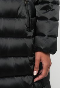 Casaco puffer preto com seções acolchoadas, exterior elegante, fecho frontal e punhos canelados. Mão visível na vestimenta.