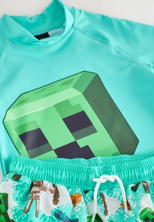 Helles blaues Rashguard mit einem grünen, pixelierten Creeper-Design auf der Vorderseite, kombiniert mit gemusterten Badeshorts, die Minecraft-Charaktere zeigen.