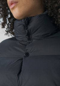 Černá péřová bunda s vysokým límcem, lesklými knoflíky a prošívanou texturou, zdůrazňující teplo a izolaci.