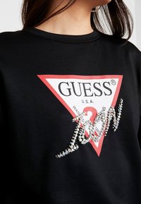 Felpa nera con una grafica triangolare rossa che presenta il logo "GUESS U.S.A." Decorata con strass luccicanti che formano la parola "GUESS."
