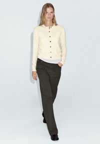 Cardigan couleur crème avec des boutons, associé à un haut blanc et un pantalon vert foncé. Le tissu semble doux, avec une silhouette ajustée et des manches longues.