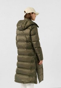 Lunga giacca a vento verde oliva con cuciture orizzontali, cappuccio e spacchi laterali. Abbinata a un cappello beige con una finitura testurizzata.