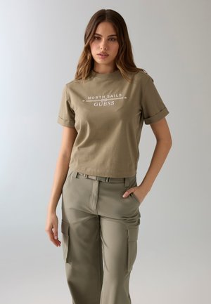Joven mujer con camiseta verde oliva "North Sails Guess" y pantalones cargo de talle alto a juego, posando sobre fondo liso.