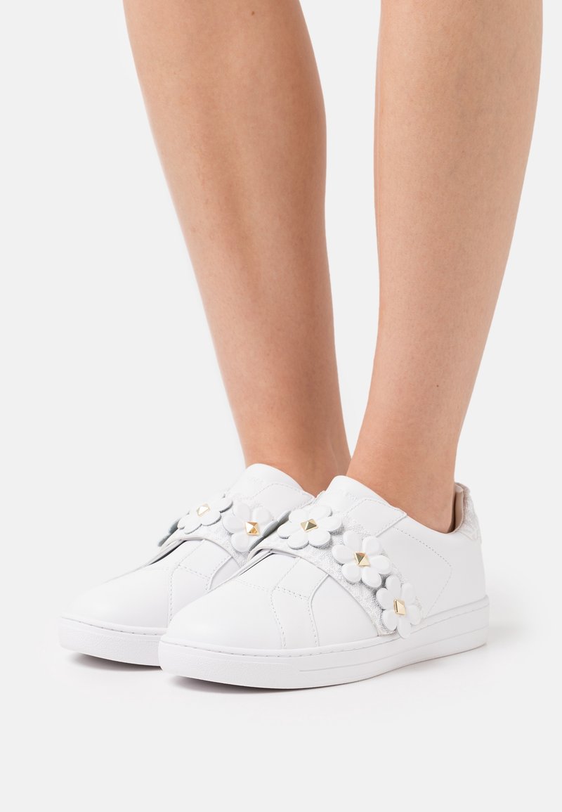 Baskets blanches sans lacets avec appliques florales blanches décoratives et clous carrés dorés, portées sur des jambes nues sur un fond clair uni.