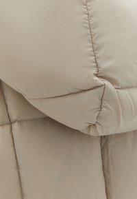Beige pufferjack met een gestikte afwerking, zachte textuur en opvallende gewatteerde delen. De stof is soepel met een subtiele glans.