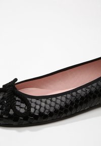 Svart balettklack med en glänsande, texturerad yta som liknar ormskinn, med en svart ribbont vid framsidan och en rosa inredning.