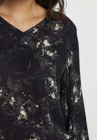 Blouse noire à col en V avec un motif éclaboussé abstrait blanc et vert foncé sur les manches longues, vue de la taille au cou.