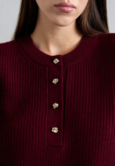 Primer piano di una donna in maglione bordeaux a coste con quattro bottoni decorativi a nodo dorato sulla patta frontale.
