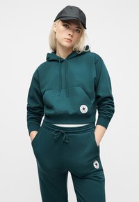 Νεαρό άτομο που φορεί πετρόλ φούτερ και φόρμα Converse με λογότυπα, μαύρο καπέλο, χέρια στις τσέπες, στέκεται μπροστά σε απλό ανοιχτό φόντο.