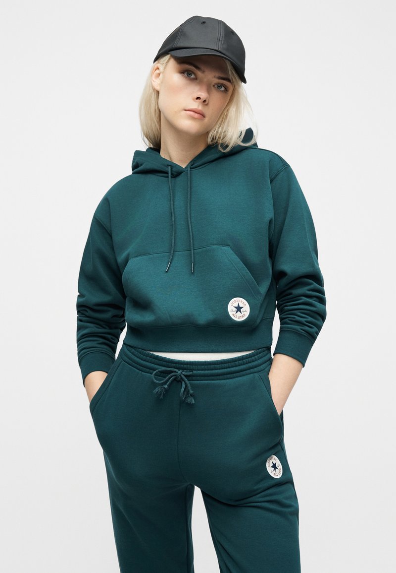 Young person wearing a teal Converse hoodie and sweatpants with logos, black cap, hands in pockets, standing against a plain light background.