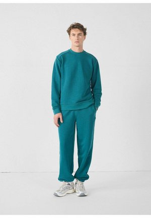 Sudadera color verde azulado y pantalones a juego en tejido suave, con mangas largas y un corte relajado. El modelo lleva zapatillas plateadas.