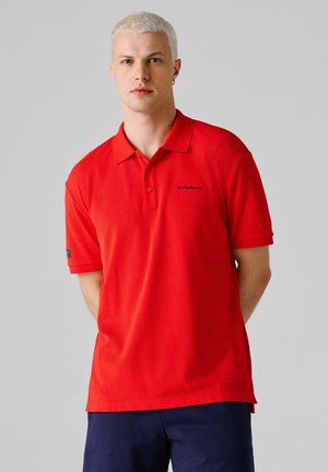 Junger Mann mit kurzen blonden Haaren, der ein rotes Poloshirt und dunkelblaue Hosen trägt, vor einem schlichten hellgrauen Hintergrund steht.