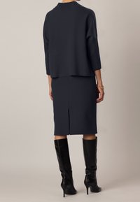 Maglione a maglia blu navy con un design a coste, abbinato a una gonna a matita coordinata con uno spacco posteriore. Indossato con stivaletti neri alti fino al ginocchio con tacco.