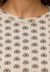 Blusa de tela color beige claro con un patrón floral repetido en marrón oscuro. Cuello redondo con detalles texturizados alrededor del cuello.