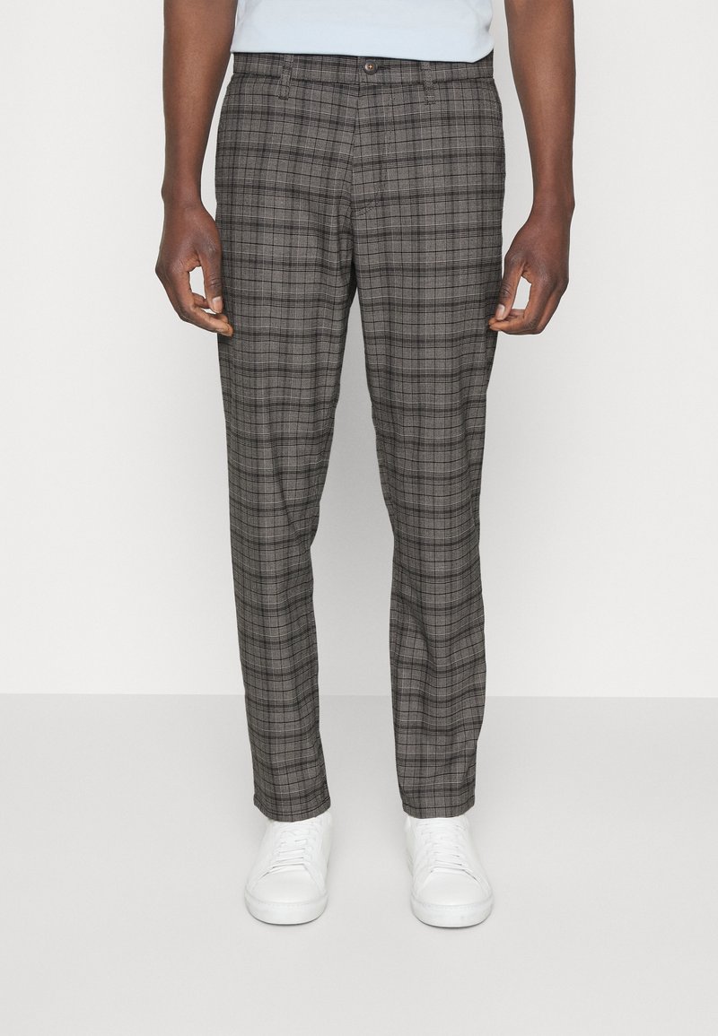 Homme portant un pantalon slim à carreaux gris et des baskets blanches, debout sur un fond blanc uni.