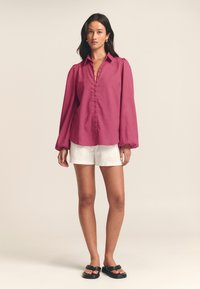 Fuchsia long-sleeve shirt with a button placket and puffed shoulders, paired with white shorts and black flat sandals on a neutral background.