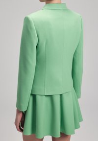 Mátově zelený blazer s přiléhavým střihem, texturovanou tkaninou a peplum sukní. Obsahuje ostrý límec a švové detaily na zadní straně.