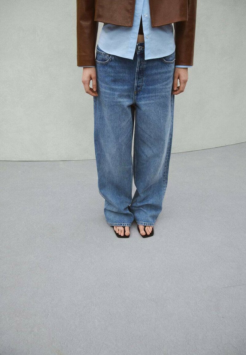 Oseba, ki nosi ohlapne modre kavbojke, rjav skrajšan suknjič, svetlo modro srajco in črne sandale s kvadratnimi prsti, stoji na svetlo sivi talni površini.