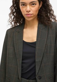 Tamsiai languota sakoma su subtiliais oranžiniais akcentais, su notch apykaklėmis, vienu mygtuku užsegama ir tekstūruotu audiniu. Dėvima ant juodo viršaus.