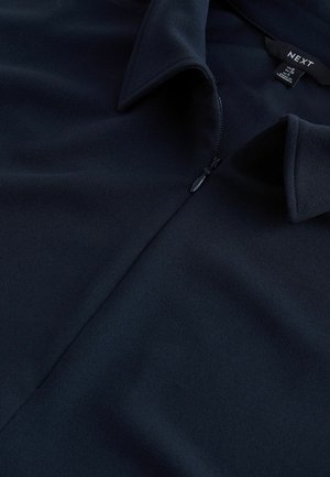 Navy zip-up blouse in a smooth, textured fabric. Features a pointed collar and a half-zip closure, with brand label visible.