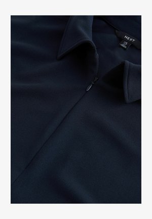 Tumši zila blūze ar rāvējslēdzēju, izgatavota no gluda, teksturēta auduma. Raksturīga ar izteiktu apkakli un pus rāvējslēdzēja aizdari, redzama arī zīmola etiķete.