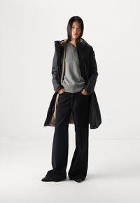 Schwarze gepolsterte Jacke mit beigem Futter, getragen über einem grauen V-Ausschnitt-Pullover, kombiniert mit hochtaillierten, schwarzen weiten Hosen. Strukturiertes Material.