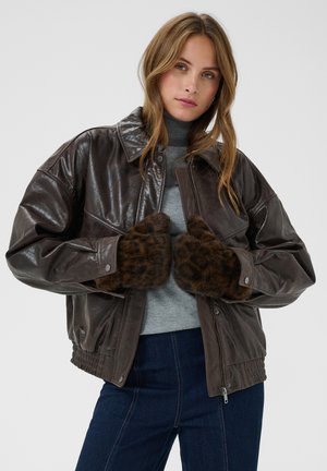 Jeune femme portant une veste en cuir marron foncé, un pull à col roulé gris, un jean bleu foncé et des gants en fourrure imprimé léopard.
