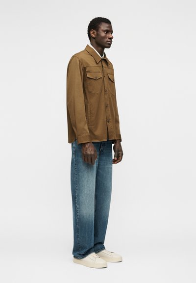 Άνδρας με καφέ πουκάμισο με τσέπες στο στήθος, φαρδιά μπλε τζιν και λευκά αθλητικά, στέκεται πλάγια σε λευκό φόντο.