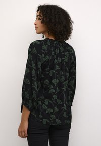 Blouse noire à motif floral avec des motifs de plantes vertes, manches longues et encolure à volants. Tissu léger, coupe décontractée, avec un ourlet arrondi.